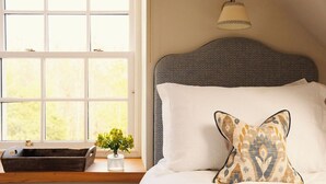 Un dormitorio con un ventanal grande, una mesita de noche con un florero, una lámpara y una almohada estampada.
