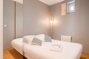 Ein ordentlich bezogenes Bett mit weißer Bettwäsche und gemusterten Kissen, ein Nachttisch mit Lampe, ein Heizkörper und ein Fenster mit Blick.