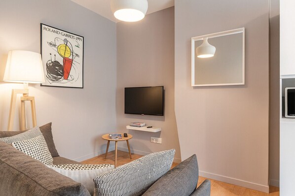 Ein modernes Wohnzimmer mit einem grauen Sofa, einem kleinen runden Tisch mit Büchern, einem Wand-Fernseher und einem gerahmten Kunstwerk an der Wand.