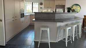 A modern kitchen with a central island, white bar stools, and a window providing natural light. - Private Family Home With Four Bedrooms (Manukau)