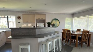 A modern kitchen with a central island, bar stools, and a dining area with wooden chairs and a table. - Private Family Home With Four Bedrooms (Manukau)