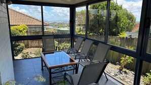 A covered patio with a table and chairs, large windows offering a view of a backyard with a wooden fence and greenery. - Private Family Home With Four Bedrooms (Manukau)
