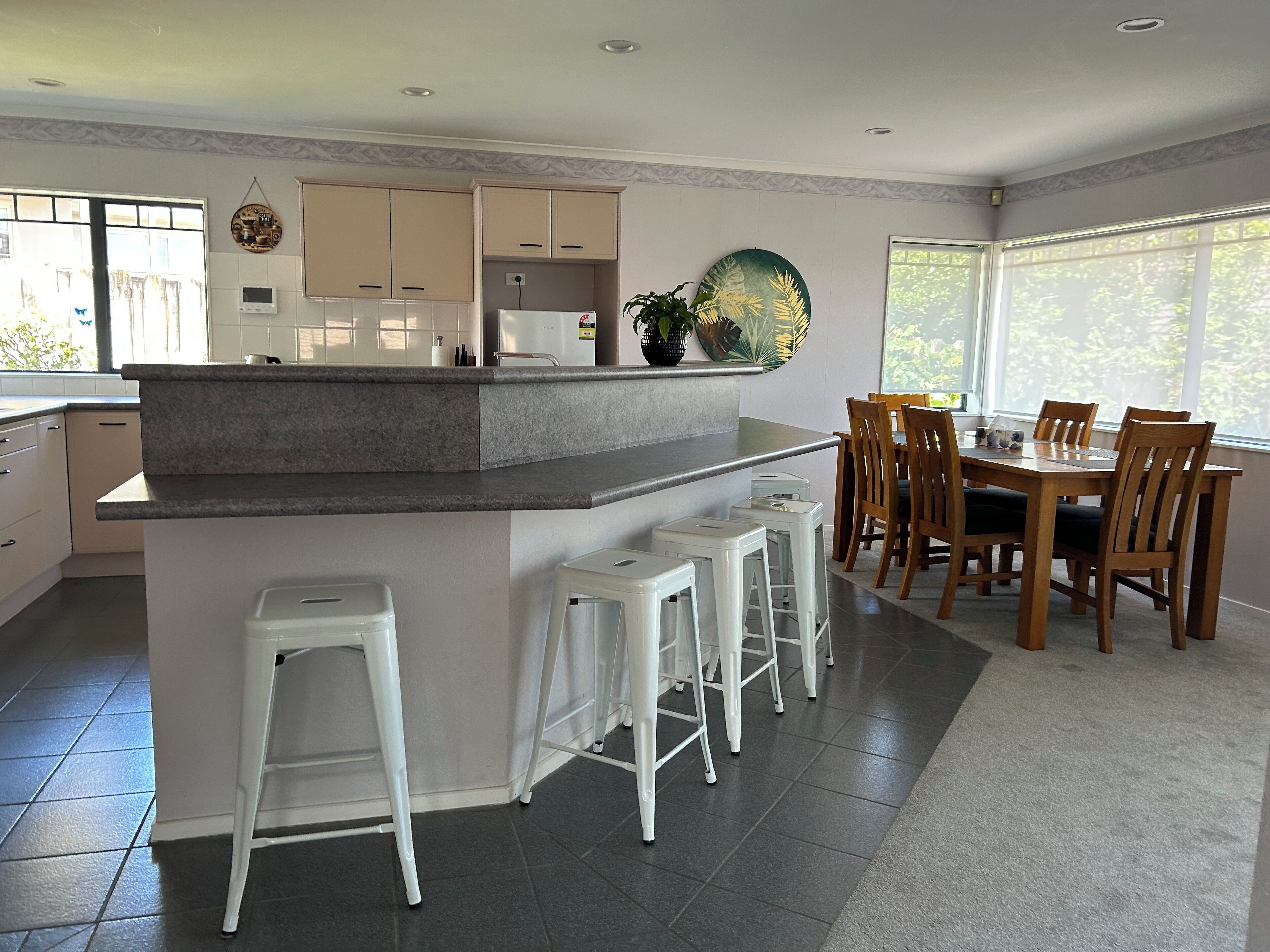 A modern kitchen with a central island, bar stools, a dining table with chairs, and large windows.