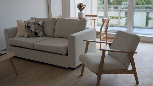 A modern living room with a beige sofa, a wooden armchair, and a small round table. - HarmoNest Rheydt Apartments 301 (Mönchengladbach)