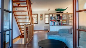 Een moderne woonkamer met een houten trap, een blauwe bank, een ronde salontafel en een boekenplank vol boeken en decoratieve objecten.