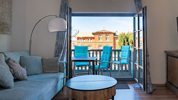Een woonkamer met een bank, een ronde houten tafel en twee blauwe stoelen. Een groot raam biedt toegang tot een balkon met uitzicht op een gebouw.