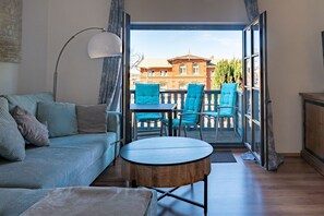 Een woonkamer met een bank, een ronde houten tafel en twee blauwe stoelen. Een groot raam biedt toegang tot een balkon met uitzicht op een gebouw.