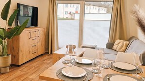 A modern living room with a wooden dining table set for four, a sofa, a TV on a wooden cabinet, and large windows with curtains. - New Tiny House - cozy, private, family-friendly! (Hessisch Lichtenau)