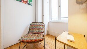 Une pièce avec un plancher en bois, une chaise dotée d’un coussin à motifs, une petite table, une lampe fixée au mur et une fenêtre avec des stores.