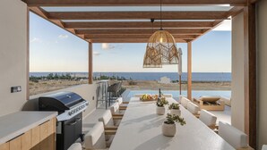 Uma área de refeições ao ar livre com vista para a praia, uma churrasqueira e um lustre suspenso.