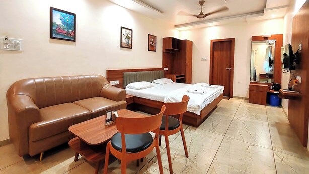 A hotel room with a bed, a sofa, a small table, and a chair. There is a ceiling fan, a mirror, and a wardrobe. - Hotel Meera Regency (Chiplun)