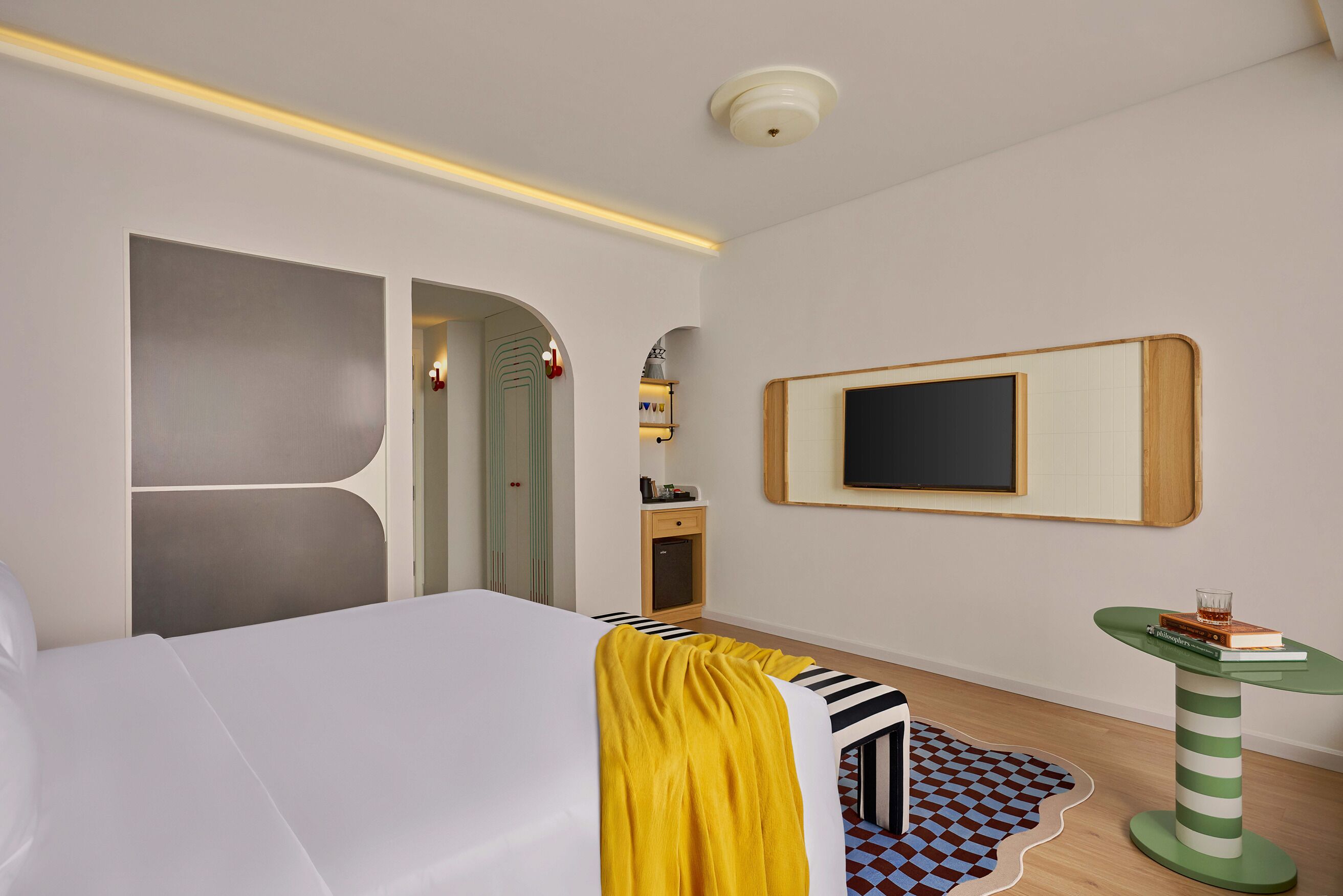 A hotel room with a bed, a desk, a chair, a small table, a television, and a door leading to another room.