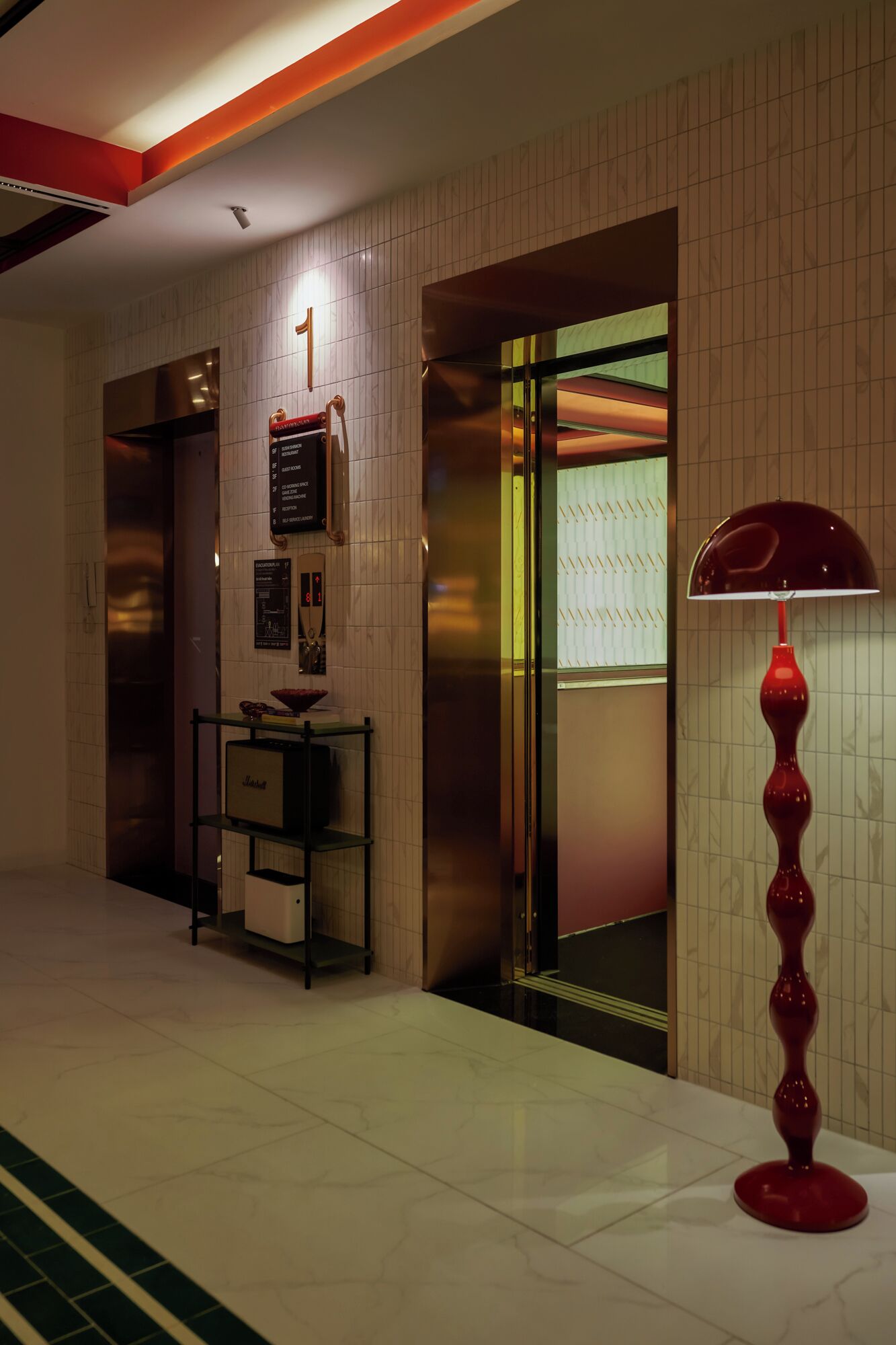 A hotel lobby with a reception desk, a red floor lamp, and a wall-mounted elevator control panel.