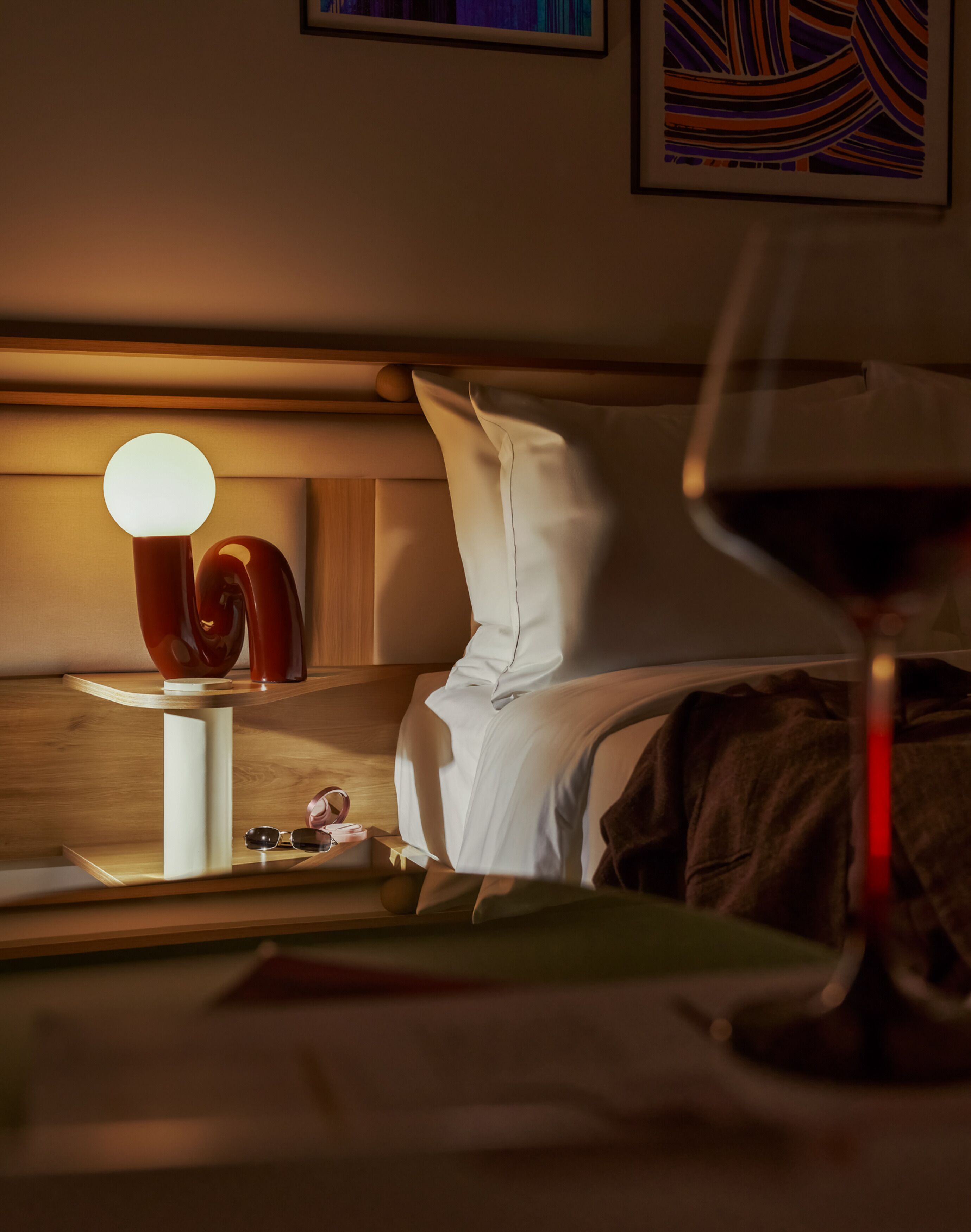 A hotel room with a bed, a bedside table with a lamp, a glass of red wine, and a framed abstract painting on the wall.