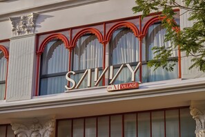 Fachada de edificio con amplios ventanales de arco y un letrero llamativo que dice 'SAVINY BY M VILLAGE'.