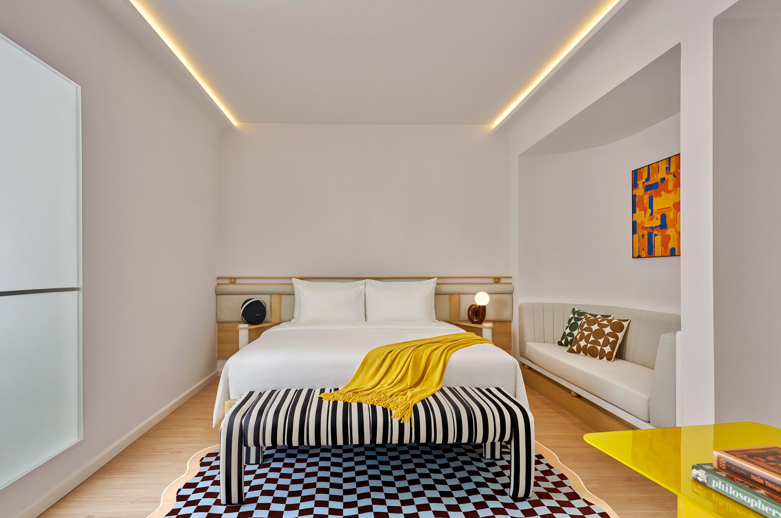 Un dormitorio moderno con una cama grande, mesitas de noche, un banco, un sofá y una pequeña mesa con libros.