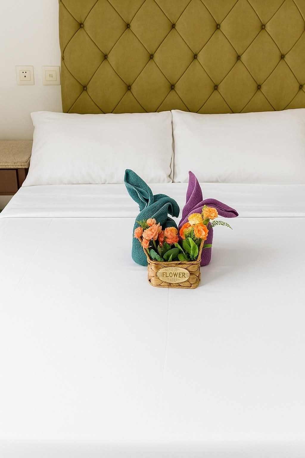 A bed with white linens, a green headboard, and a small wicker basket with a label "FLOWER" containing colorful flowers.
