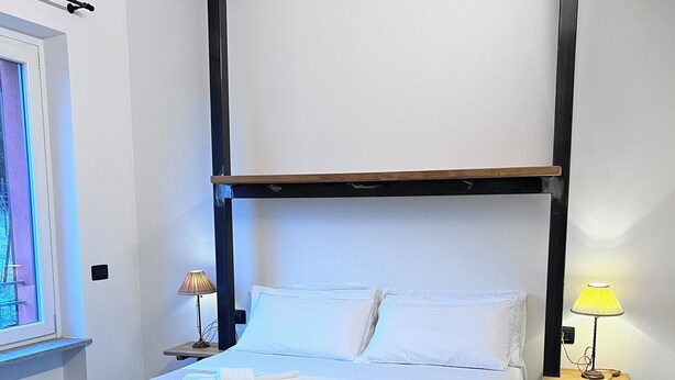 A neatly made bed with a wooden headboard, a bedside table with a lamp, and a window with a view of greenery. - App. Red (Borgonovo)