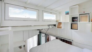 Et værelse med køjeseng, to vinduer, en hylde med et billede i ramme og en lille hængende lampe.