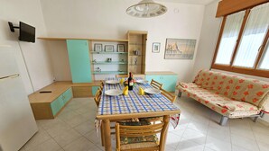 A dining area with a checkered tablecloth, wooden chairs, a sofa, and a cabinet with shelves. - Pleasant apartment in Rosolina Mare with balcony (Rosolina Mare (RO))