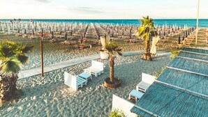 A beach with rows of white lounge chairs and umbrellas, palm trees, and a clear view of the ocean. - Pleasant apartment in Rosolina Mare with balcony (Rosolina Mare (RO))