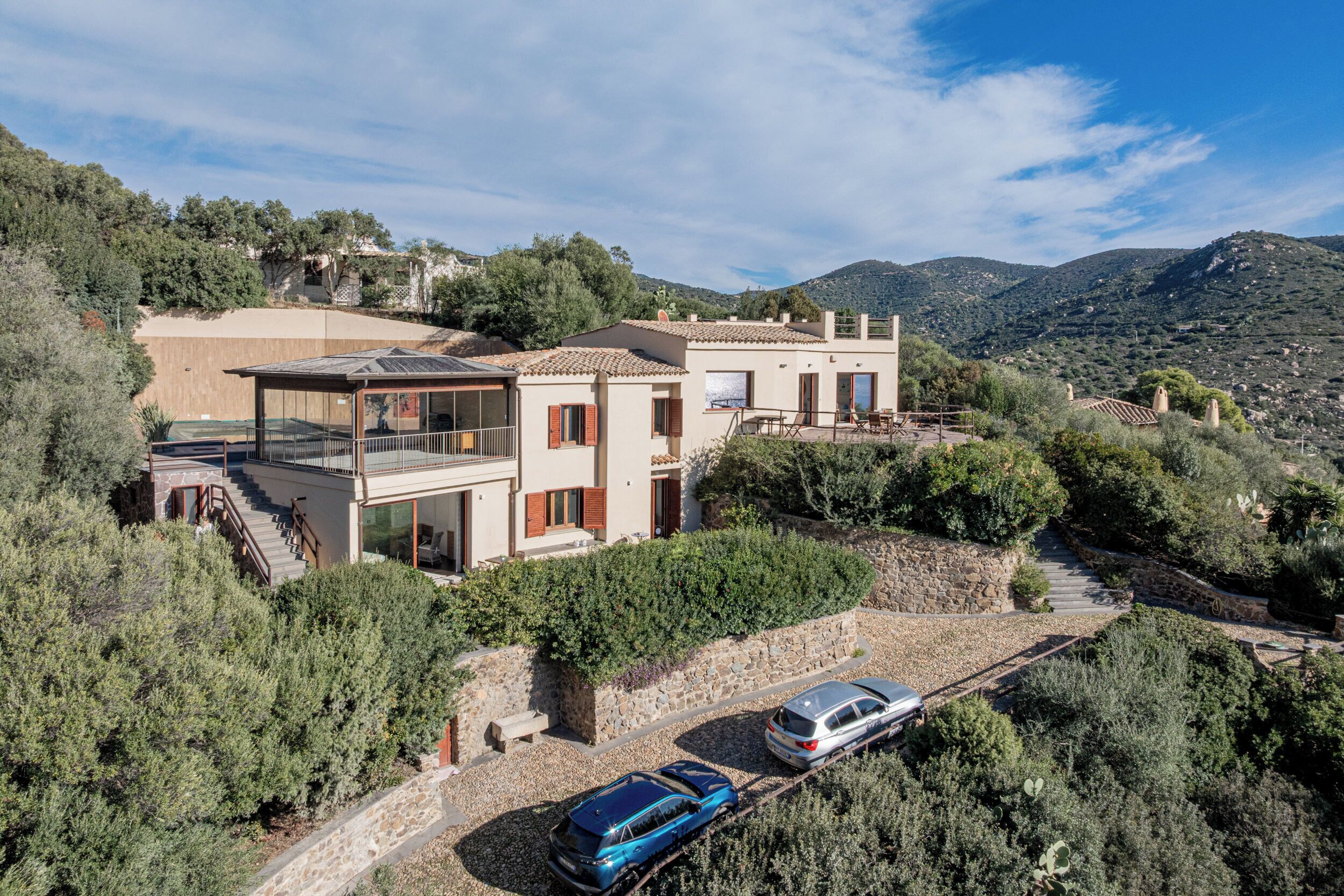 Une maison moderne à deux étages, dotée d’un balcon, avec une voiture garée devant et un paysage montagneux en arrière-plan.