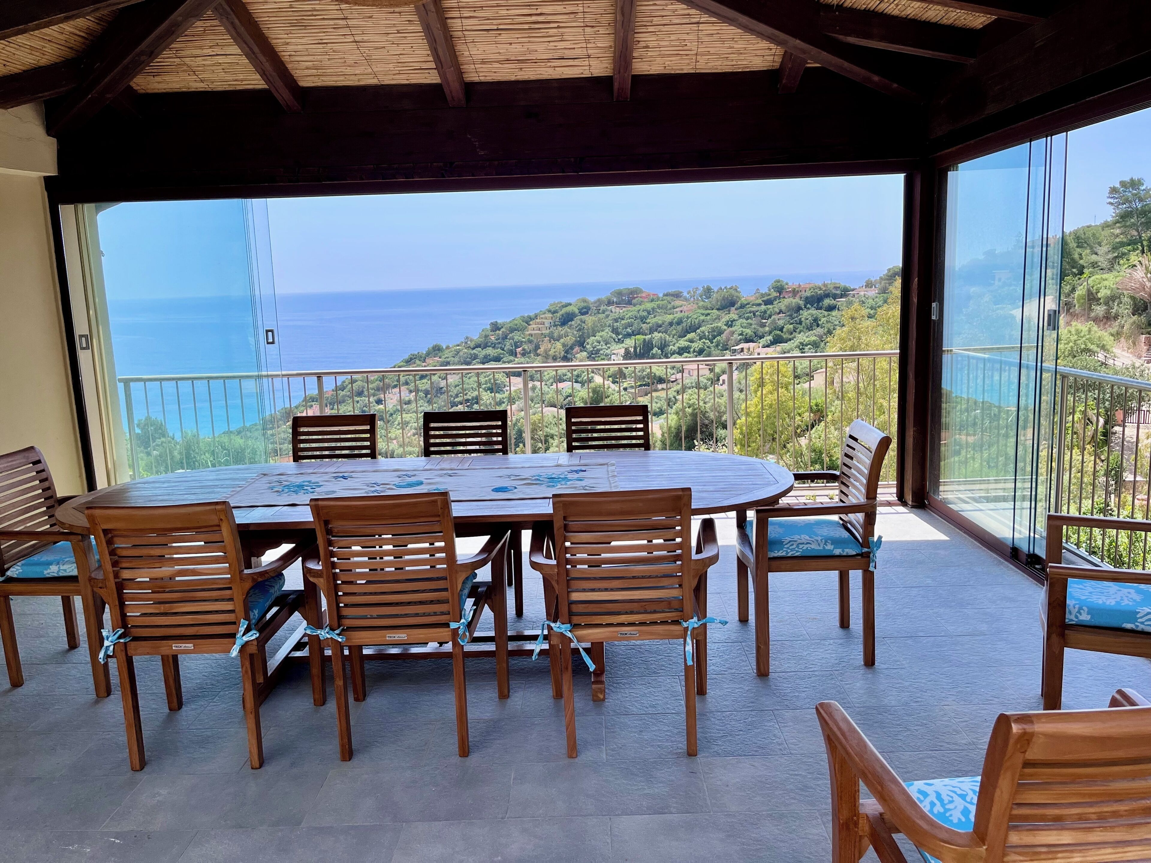 Un espacio de comedor al aire libre cubierto, con mesa y sillas, con vistas panorámicas al mar y a las colinas.