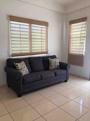 Um sofá escuro com almofadas estampadas em um quarto com piso de azulejos e duas janelas com persianas.