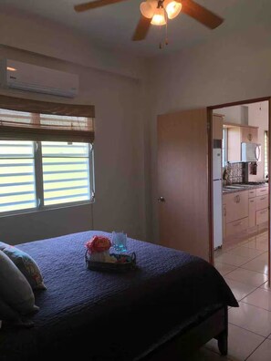 A bedroom with a bed, a window with blinds, a ceiling fan, and a view into the kitchen. - Apartments Pepe's View - D (Bajío)