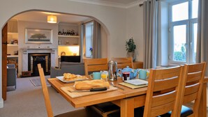 En matplats med ett träbord dukat för frukost, en öppen spis i bakgrunden och utsikt över naturen genom ett fönster.