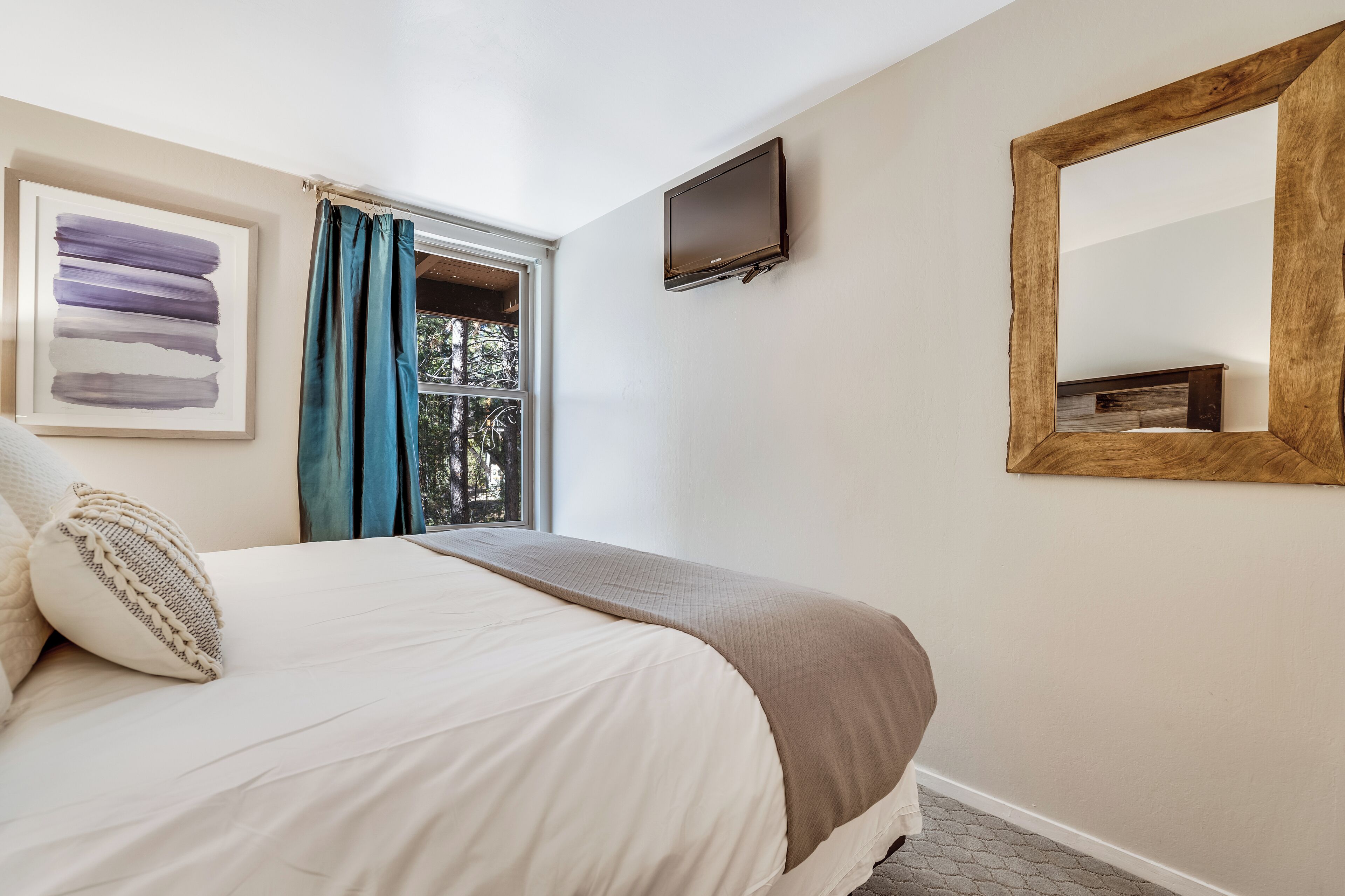 A bedroom with a bed, a window with curtains, a mounted TV, and a mirror on the wall.