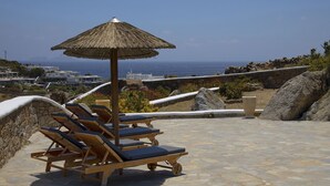 A sunlit outdoor lounge area with wooden lounge chairs and a thatched umbrella, overlooking a coastal town and the sea. - My Rental Homes - Villa Cassandra, less than 2 km from the beach with pool (Mykonos)