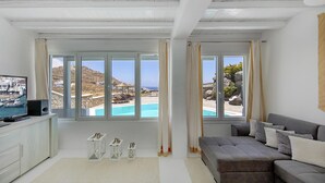 A modern living room with large windows, a grey sofa, a flat-screen TV, and a pool view. - My Rental Homes - Villa Cassandra, less than 2 km from the beach with pool (Mykonos)