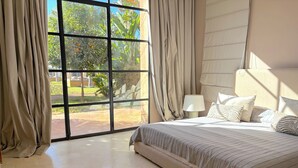 Quarto com uma grande janela, uma cama com roupa de cama listrada e uma mesa de cabeceira com abajur.