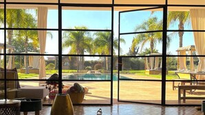 Quarto com uma grande janela com vista para uma piscina e palmeiras, com uma mesa de madeira com livros e objetos decorativos.