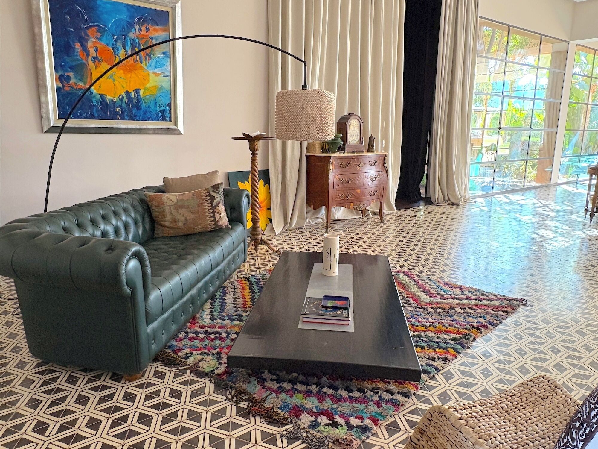 Un salón con un sofá verde, una mesa de centro con libros, una alfombra colorida y un gran ventanal con cortinas.