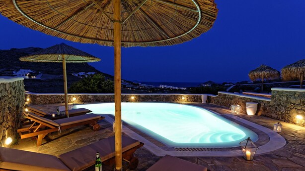 A pool area with lounge chairs and umbrellas, surrounded by stone walls and overlooking a coastal landscape. - Panoramic Villa Fos, in Mykonos, in Kalafatis area, with private pool, 4 bedrooms, 10 sleeps (Mykonos)