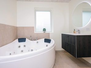 Un baño moderno con una bañera independiente, una pequeña ventana con una planta, un espejo redondo y un mueble de tocador de madera oscura.