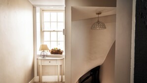 Un couloir étroit avec un mur blanc, une fenêtre, une petite table avec une lampe et un luminaire décoratif suspendu au plafond.