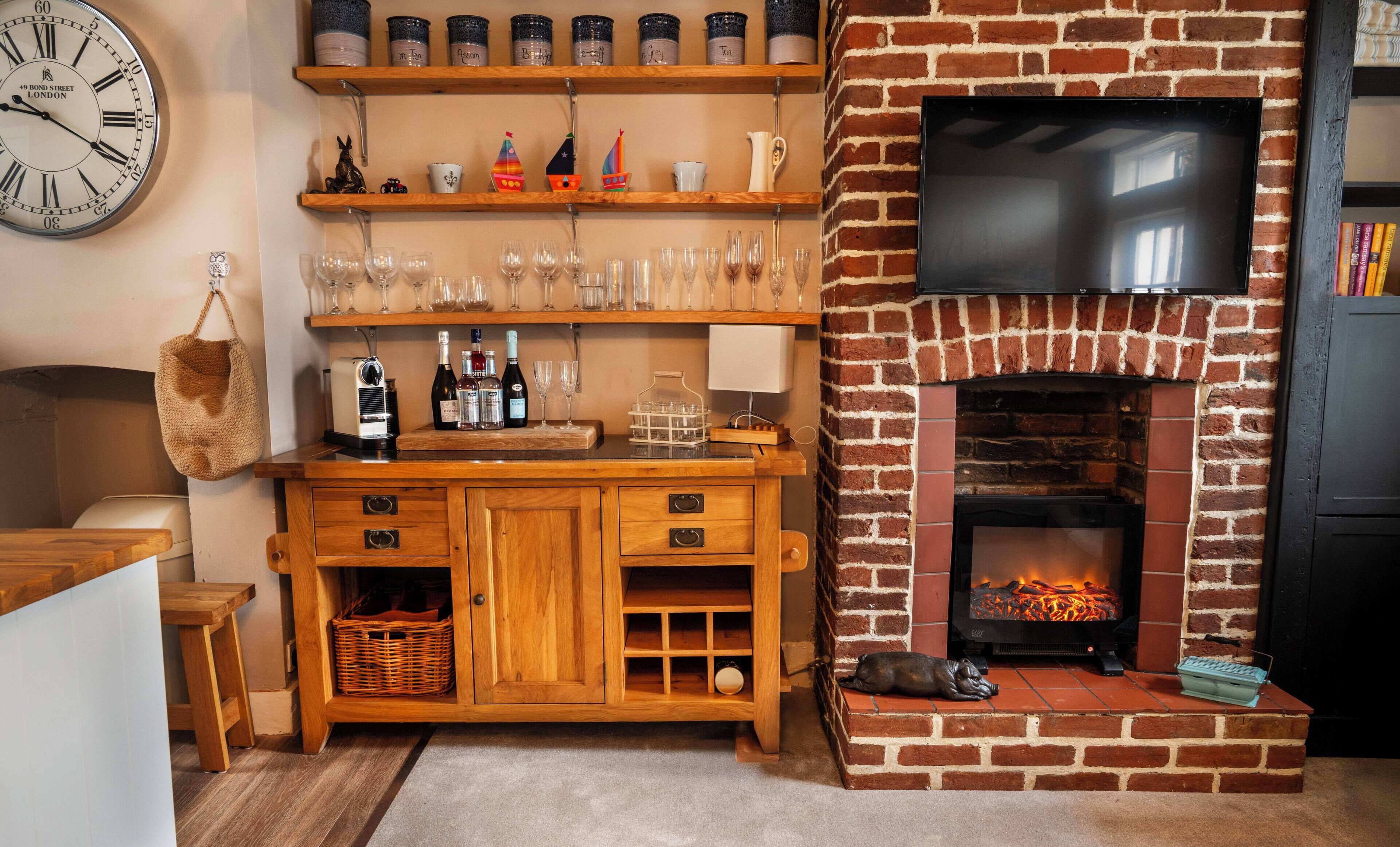 A cozy living room with a brick fireplace, wooden shelves with decorative items, a clock, and a television.