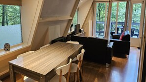 A modern kitchen with a wooden table and white chairs, a black sofa, and a view of outdoor seating through large windows. - Serene Vacation Chalet in Durbuy (Durbuy)