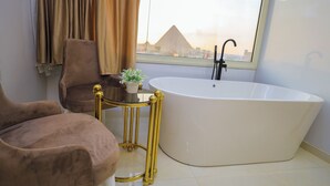 Un baño moderno con una bañera independiente, un tocador con marco dorado, un sillón marrón y vistas a las pirámides.
