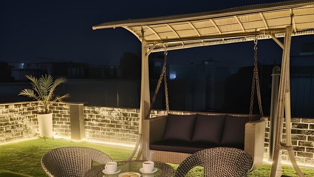 Un espacio de estar al aire libre con sillas de mimbre y una mesa, bajo una pérgola cubierta con luces colgantes, frente a un muro de ladrillo y un césped.