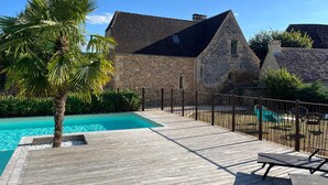A wooden deck with a swimming pool, stone buildings, and a palm tree. - Valley View – Large house for 10 – Pool (Les Eyzies-de-Tayac-Sireuil)