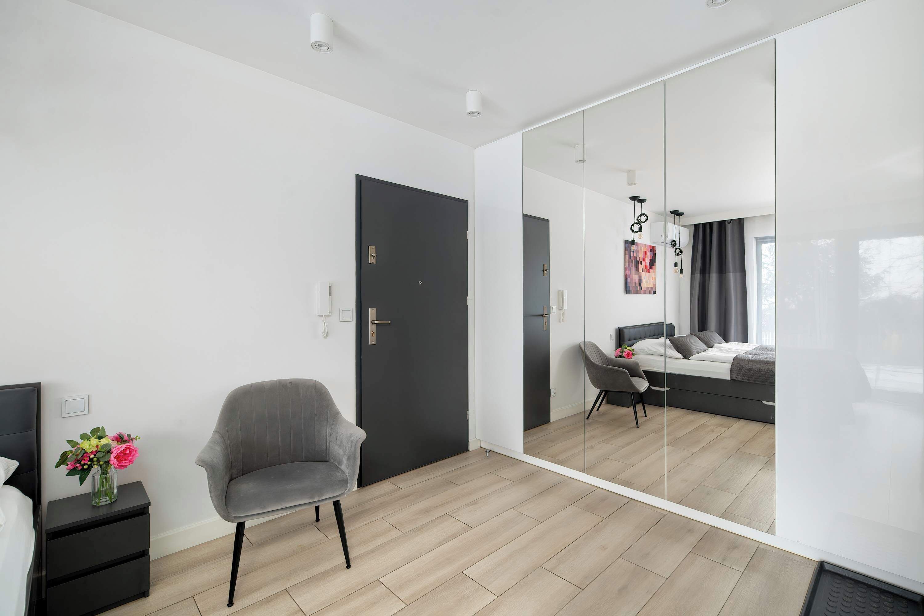 Een moderne slaapkamer met een grote spiegel, een grijze stoel, een nachtkastje met een bloemenvaas en een bed met een grijze hoofdbord.