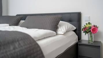 Un llit ben fet amb roba de llit gris i una almoina blanca, una petita taula de nit amb un vas de flors, i una paret sense decoració amb un interruptor.