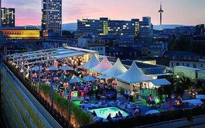 Evento en la azotea con carpas y piscina, rodeado de edificios de la ciudad y una torre.
