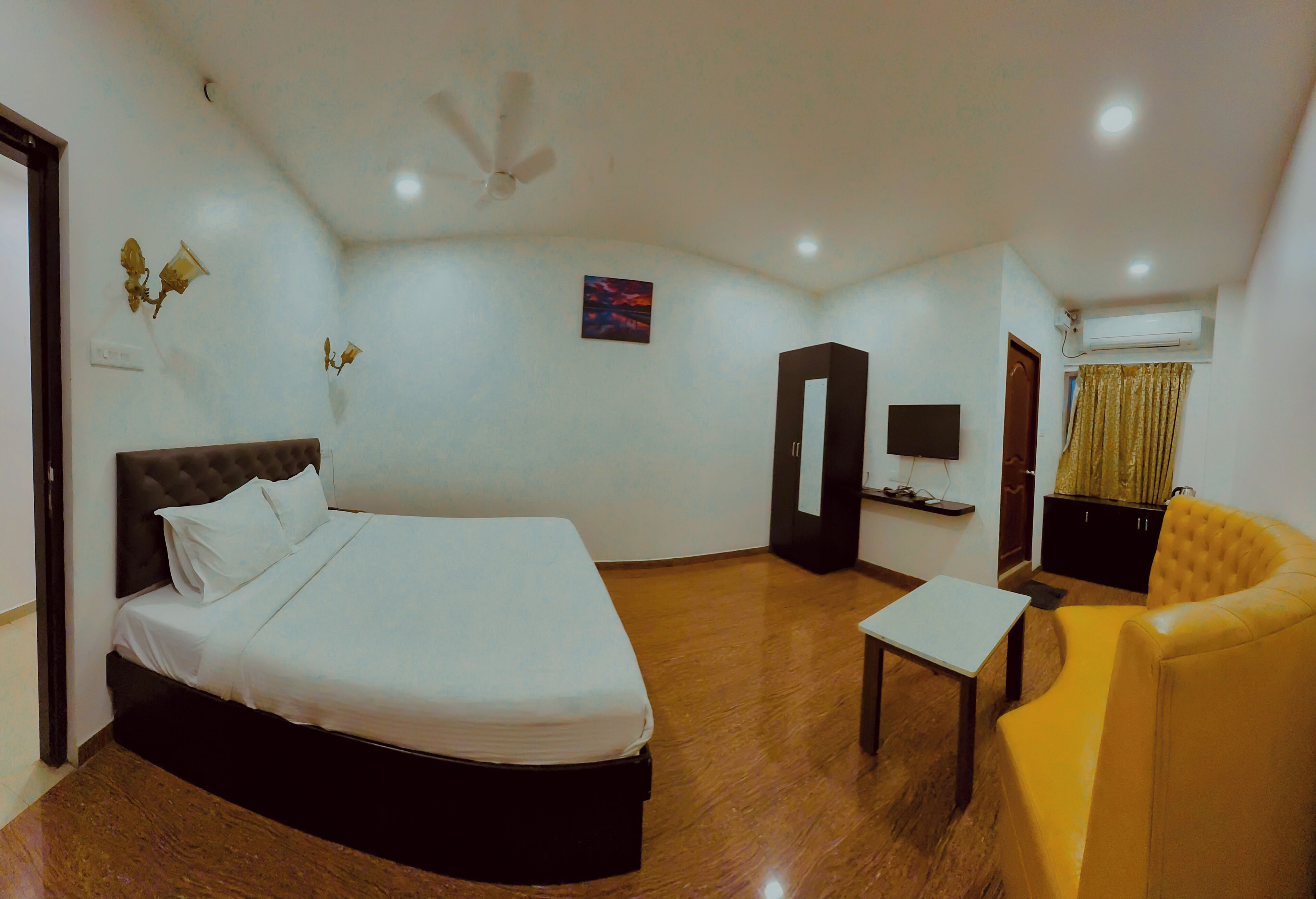 A hotel room with a bed, a small table, a yellow armchair, a wardrobe, a television, and an air conditioning unit.