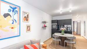 A modern living room with a sofa, a dining table, and chairs. There is a kitchen in the background with cabinets and a refrigerator. - 2-room apartment in Charlottenburg-Wilmersdorf (Berlin)