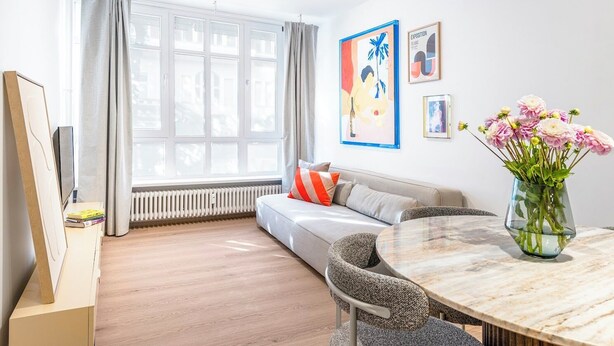 A modern living room with a sofa, a dining table, and a large window with curtains. There are framed artworks on the wall and a vase with flowers on the table. - 2-room apartment in Charlottenburg-Wilmersdorf (Berlin)